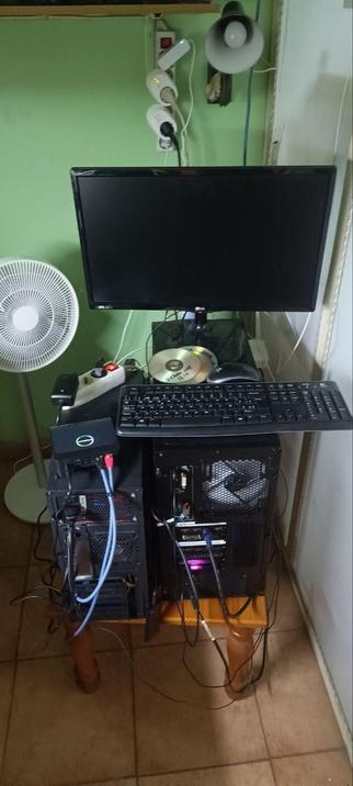 Foto en la que se ven dos servidores negros sobre un banco, con un monitor y un teclado encima, todo lleno de cables. Detrás se puede ver un ventilador, enchufes y una lampara de pared.