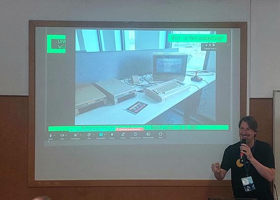 The image shows a presentation taking place in a room with a large projection screen. The screen displays a photograph of an old computer setup, including a monitor, keyboard, and other peripherals on a desk. The photograph is labeled with the text "What is Retrocomputing?" at the top. The screen also shows a green bar with the text "UNI WU" in the top left corner and "DH2025 Listener | 18 JUL 2023" at the bottom. A person is standing to the right of the screen, holding a microphone and wearing a black shirt with a lanyard and badge. The room has a simple, functional design with a wooden panel on the wall and a window with blinds in the background.
