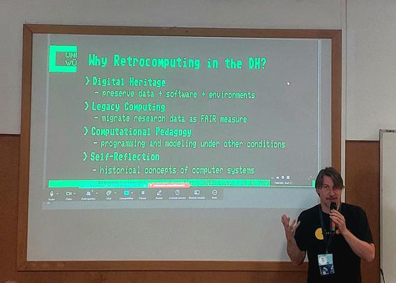 The image shows a presentation slide titled "Why Retrocomputing in the DH?" displayed on a large screen. The slide is divided into four main sections, each with a subheading and bullet points. The sections are:
Digital Heritage - preserve data + software + environments
Legacy Computing - migrate research data as FAIR measure
Computational Pedagogy - programming and modeling under other conditions
Self-Reflection - historical concepts of computer systems
The slide is projected with a green font on a white background, and the date "18 JUL 2025" is visible at the bottom, along with the location "Lisbon." The presenter, a man wearing a black t-shirt with a yellow circle on it and a lanyard with an ID badge, is standing to the right of the screen, holding a microphone and gesturing with his left hand. The background is a plain wall with a wooden panel on the left side.