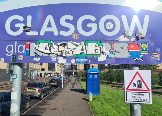 A sign with a picture of a police box on it with the words Caution Time Lords Crossing Ahead. In the background is a police box.