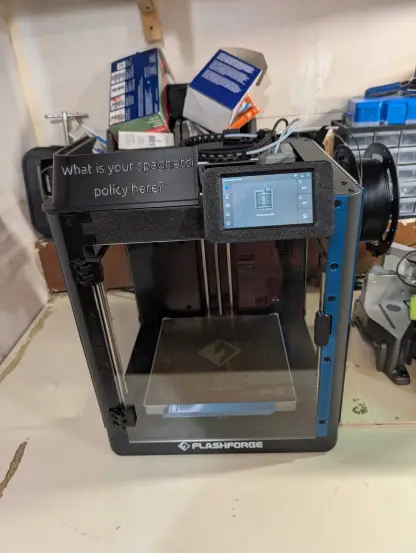 a black 3d printer sitting on a white desk. the 3d printer has silver letting on the top left of the enclosure that reads: "what is your spaghetti policy here?"