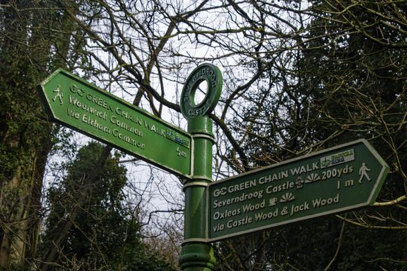 A metal post, painted in green has a donut on top with the words Severndroog Castle. There are two pointers, in green with white letters pointing to several destinations along the Green Chain Walk.
