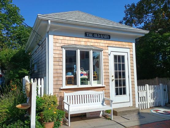 A photo of a small cottage with newly replaced cedar shingles, a bay window, and a french door there is a white bench below the window on the sidewalk and a picket fence on either side to the left are a few flower pots with plants. below the roofline is a sign that reads, confusingly, "The Old Baby".