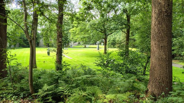op de voorgrond een aantal bomen en laag struikgewas. Tussen de bomen door is een heldergroen grasveld te zien met daarop verspreid allemaal verschillende beelden