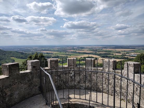 Im Vordergrund Zinnen einer Burg mit Aussicht auf die weite Hohenlohe. Darüber ein wolkenverhangener Himmel.