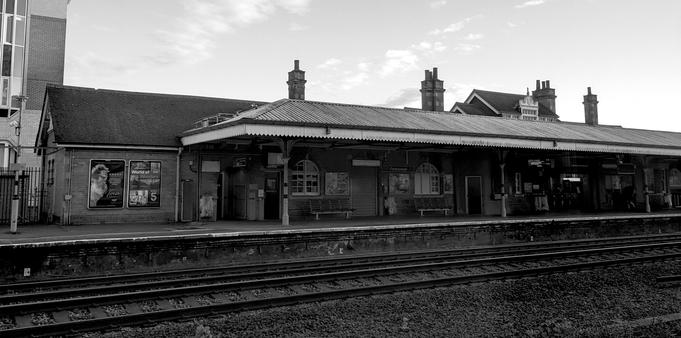 A black and white photo of a train station platform with benches, posters, and closed shutters; train tracks run parallel in the foreground.

Ein Schwarz-Weiss-Foto eines Bahnsteigs mit Baenken, Plakaten und geschlossenen Fensterlaeden; im Vordergrund verlaufen parallel verlaufende Bahngleise.