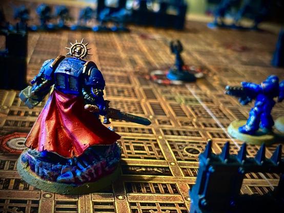 Nahaufnahme einer bemalten Warhammer-40k-Figur eines Ultramarine-Captains mit rotem Umhang und goldener Rückenverzierung, der auf einem detaillierten Spielfeld steht. Im Hintergrund weitere Space Marines, Miniaturen und Geländestücke.