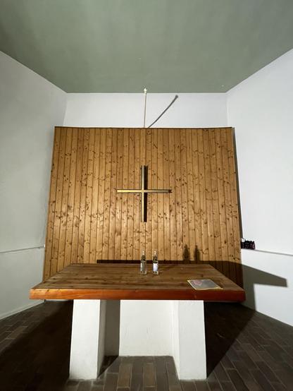 A minimalistic room featuring a wooden wall with a cross, a wooden table with two bottles, and a sheet of paper on it. The floor is made of wooden tiles, and the ceiling is painted green. Apparently a small church building