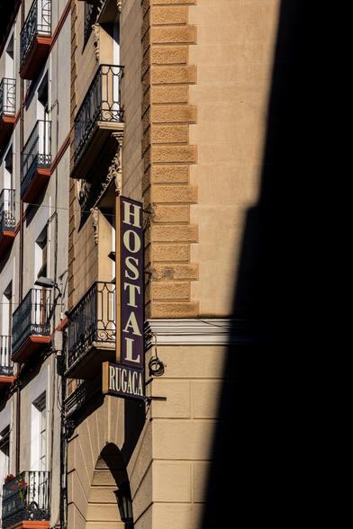 Facade of an urban building with wrought iron balconies and a vertical sign reading "HOSTAL RUGACA", partially lit by sunlight with a deep shadow on the right side.