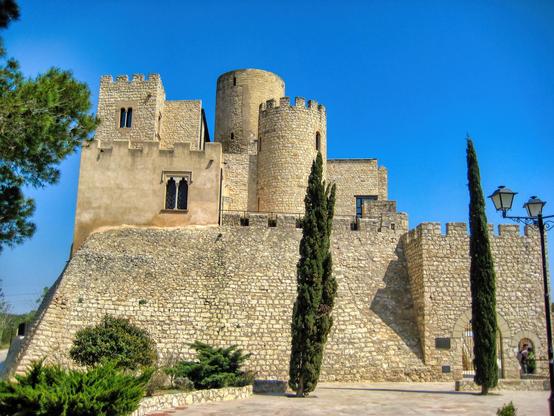 La imatge mostra un castell de pedra, amb murs de pedra robustes i torres cilíndriques. L'estil arquitectònic sembla medieval o renaixentista, amb una combinació de pedra de tons clars i beix i algunes zones d'ocre. Hi ha arbres de fulla perenne, possiblement xiprers, plantats davant del castell i també vegetació baixa prop de les muralles. El cel és blau clar i clar, contrastant amb els colors terrosos de l'edifici. A la cantonada dreta es veu un pal de llum d'estil antic. En conjunt, la imatge projecta un sentit de la història, la solidesa i la serenitat.