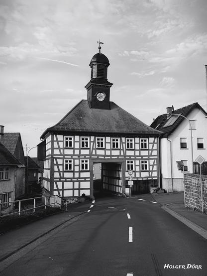 Schwarz-Weiß-Foto des barocken Torhauses in Lich-Oberbessingen. Das Fachwerkhaus mit Uhrturm steht frontal im Bild und besitzt eine große Durchfahrt, durch die eine Straße hindurchführt. Der Kontrast betont die Struktur des Fachwerks, das Dach wirkt dunkel und massiv, der Himmel ist bewölkt mit weichen Wolken. Die Aufnahme vermittelt eine ruhige, zeitlose Atmosphäre.