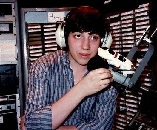 Dark haired college student wearing headphones and a striped shirt is talking into a microphone. Carts of music are set up behind him and a small Pound Puppy is sitting atop the microphone.