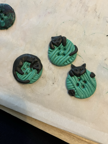 Three 1” diameter circular buttons made to look like sea green balls of yarn, with black cats partially curled around them.