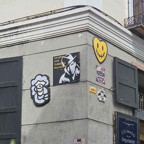 Esquina de un edificio en el barrio de Malasaña, Madrid, decorada con arte urbano. En la pared gris se observan varias piezas de arte callejero: una especie de arte abstracto con un ojo en el centro formando una cara, una imagen en blanco y negro de Humphrey Bogart con el texto "SIEMPRE NOS QUEDARÁ MALASAÑA", un corazón amarillo con una sonrisa cosida, un personaje pixelado en tonos rosas, una placa amarilla y la cara caricaturesca de un mapache sonriente.