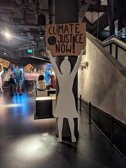 A cut-out silhouette of a child holds a "CLIMATE JUSTICE NOW!" sign against a dark pillar in a museum exhibition. The cut-out is white, depicting a figure with a bobble hat, raising the brown cardboard sign above its head. In the background, other exhibition elements are visible, including figures holding signs with German text like "DIE UHR TICKT" (The clock is ticking) and "UNSERE ZUKUNFT SCHMILZT MIT" (Our future is melting too). Several blurred people are also present in the dimly lit space with overhead lighting. A dark staircase is on the right.