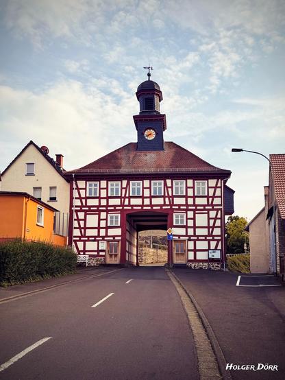 Frontalaufnahme des barocken Torhauses in Lich-Oberbessingen von der Nordostseite. Das rot-weiße Fachwerkhaus mit Uhrturm steht mittig im Bild, durch die große offene Durchfahrt ist eine helle Steinmauer sichtbar. Der Himmel ist leicht bewölkt, das Licht gleichmäßig. Die Szene wirkt ruhig und klar strukturiert.
