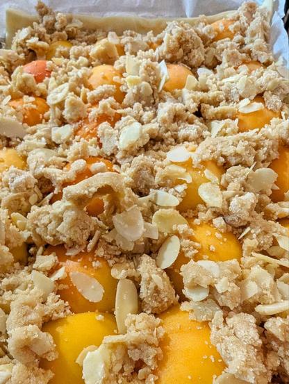 Kleines Backblech mit ausgerolltem Hefeteig, der kaum sichtbar ist, unter den Kuppen der darauf liegengen Aprikosehälften, die unter zimtigen Streuseln begraben sind, die mit Mandelblättchen angereichert sind. (ungebacken)