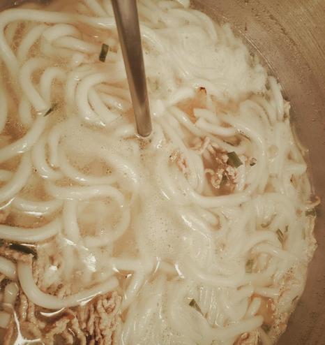 A photo of banh canh noodle soup being stirred in a metal pot with a metal ladle. The soup consists of thick, rounded, and white udon noodles, ground meat, scallions, and a light-colored broth with bubbles. The ladle is inserted into the center of the pot