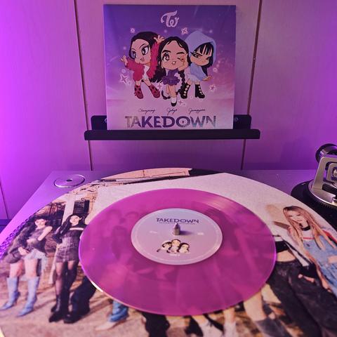 A transparent violet vinyl record sits on a turntable. Behind the turntable, a vinyl album outer sleeve is displayed. The front cover shows chibi versions of TWICE members Chaeyoung, Jihyo, and Jeongyeon.