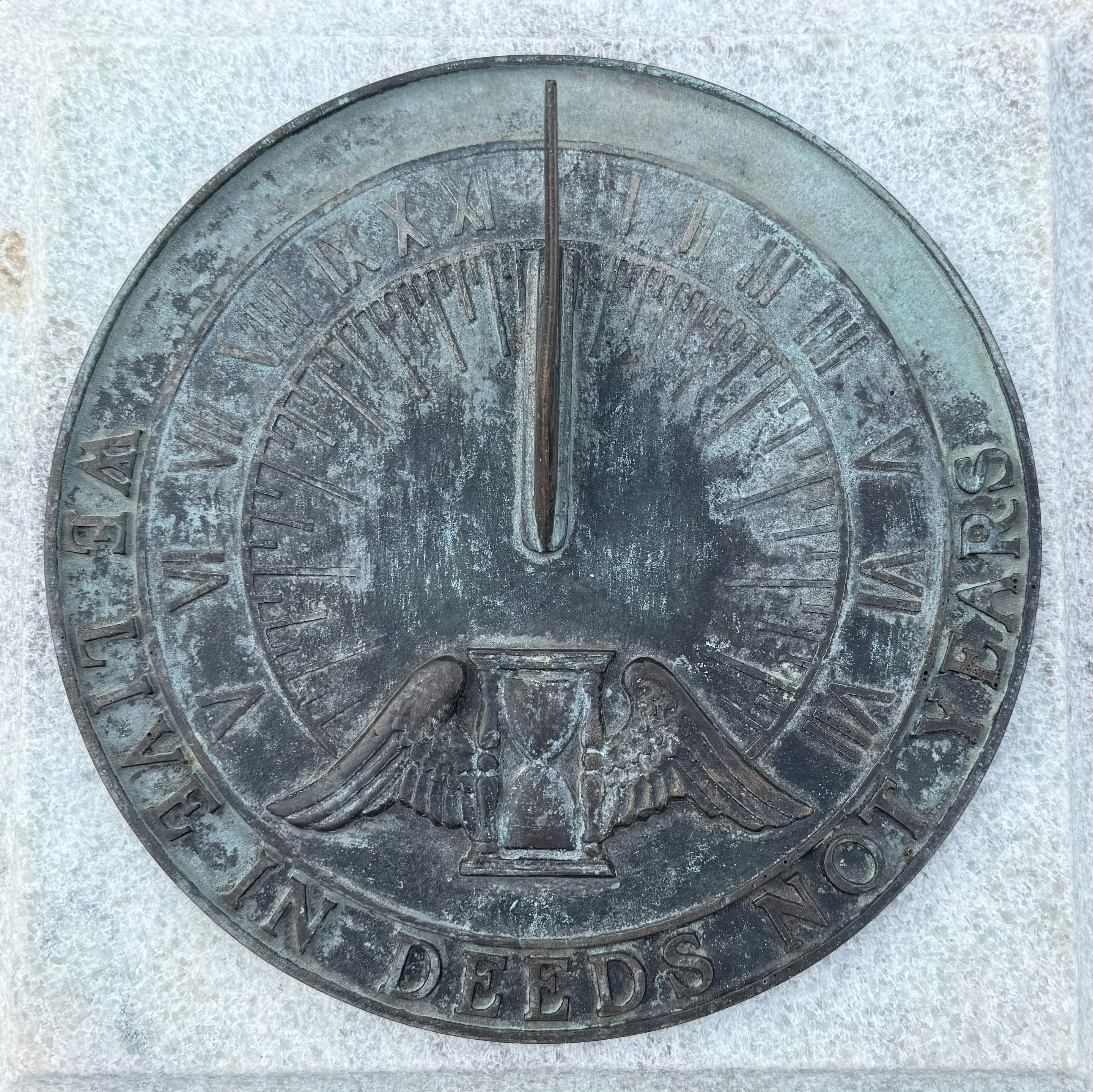 A bronze sundial on a piece of granite. The bronze is weathered and green with oxidation. An hourglass with wings appears beneath the gnomon, and there are Roman numeral markings for the hours. In a semicircle outside of the dial is the inscription WE LIVE IN DEEDS NOT YEARS.