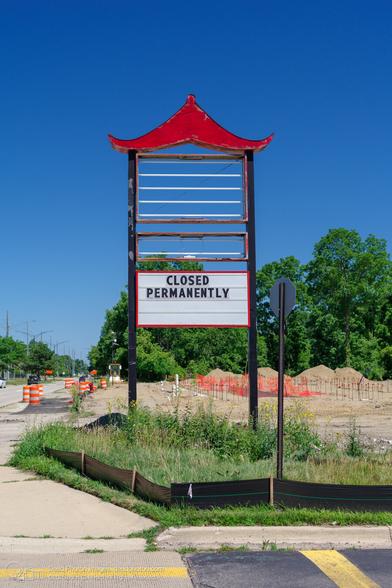 A sign with a red pagoda-style roof reads "CLOSED PERMANENTLY" in black letters. The sign is in front of a construction site with orange safety fencing. The sky is cloudless and blue.