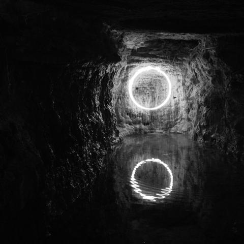 View of a small cave at night, with its lake reflecting a circle-like neon light fixed to the back wall. (Version françæ) Vue d’æn petix grotte læ nuit, avec saon lac, dans læquæl se reflète æn éclairage al néon en forme de cercle fixæ sur læ paroi dal fond.