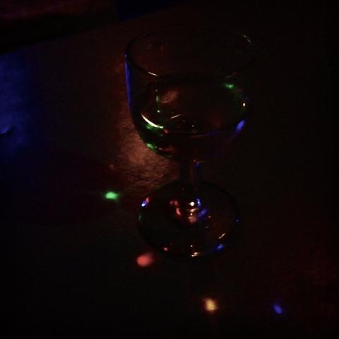 A small, round glass with a stem reflects the light from a multicoloured garland, creating small, coloured dots all around. (Version françæ) Petix verre ronx à pied reflétant læ lumière d’æn guirlande multicolore, ce qui crée des petiz points coloræs tout autour.