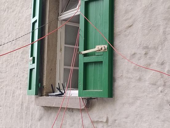 Ein Fenster mit offenen Fensterläden und offenen oberlichtern aus denen 7 Kabel hängen und aus dem Bild heraus führen. Außen auf der Fensterbank ein Router mit vielen Antennen.