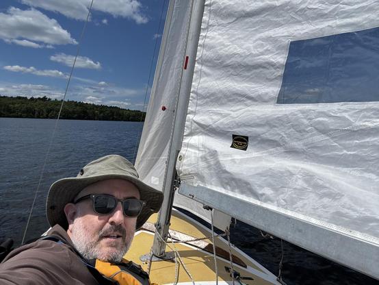 A man wearing sunglasses and a hat on a small sailboat on a lake with blue skies