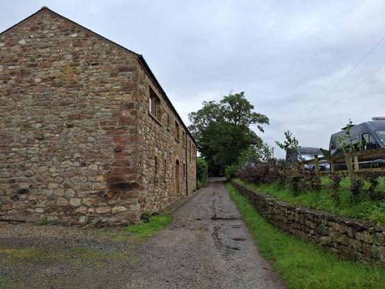 Picture of a stone farm building, with a small road next to it. The road leads in the distance parallel to the building, and on the right of it is a field where we can see two campervans (neither of which is mine!)