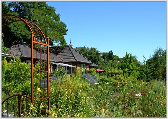 Das Bild zeigt einen Garten, den man durch einen braunen Metallbogen betritt. Im Hintergrund stehen mehrere kleine Häuser mit Ziegeldächern. Der Garten ist voller blühender Pflanzen und Sträucher. Der Himmel ist klar und blau.