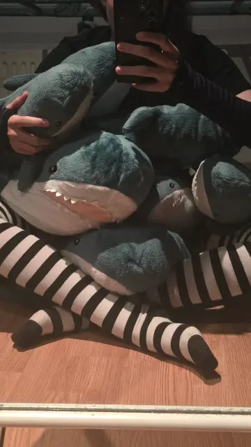 Me sitting cross legged on a Blahaj with a ton of other blahajs on my lap. I am wearing black and white thigh highs with a skirt, and arm warmers