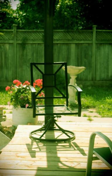 A medium shot of a metal swivel deck chair without a cushion sitting on the wooden deck, a metal post immediately behind it; slightly further back is a large white deck planter full of flowering plants.  In the background is a birdbath and a wooden fence. The atmosphere is one of stillness on a warm summer afternoon.