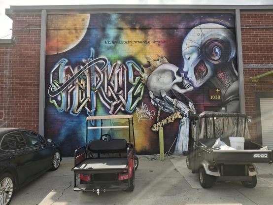 A large mural on a brick wall in graffiti style featuring a sun-like orb (hail it) and rainbow colored clouds with an android holding a human skull with a crown up to the android's artificial face.