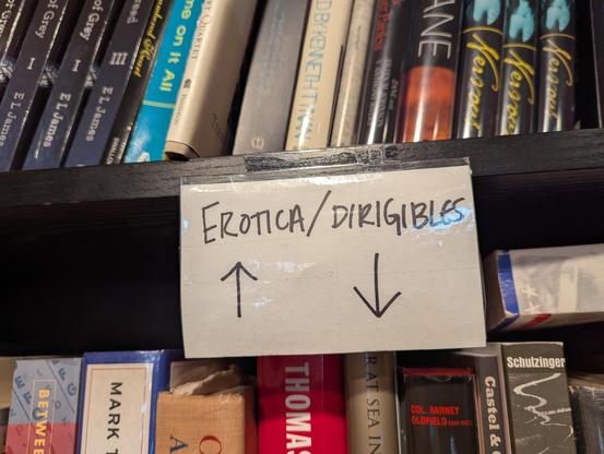 A sign on a bookshelf reading "Erotica / Dirigibles", with arrows indicating that the former is on the shelf above, and the later on the shelf below.