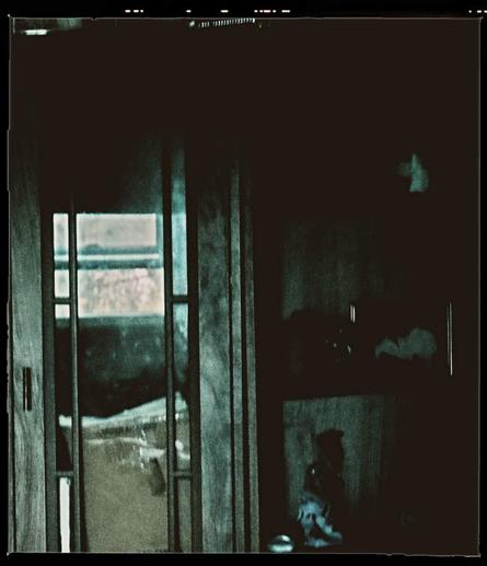 A reflection of a window appears in the glass pane of a cabinet door. Within the cabinet, all that can be seen is what appears to be a cardboard box. To the right of the door is shelving, that is dark and blurred. Vague forms of some ornaments can be seen.