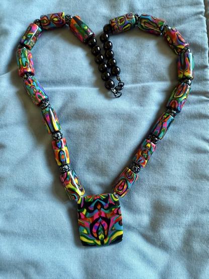 Rainbow colors and black, in random patterns, on a pendant and on 16 cylindrical beads. The clay beads alternate with small, spherical black and white glass beads. Small black onyx beads, six per side, fill the top to make the necklace 22 inches long.