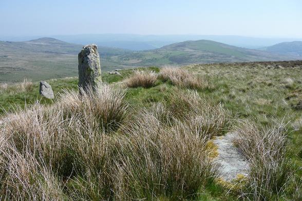 Three stones of a possible stone row on a grassy mountain ridge. The stone on the left is a short stump. The middle stone is a 6 foot tall pillar. The stone on the right lies fallen and is partly buried in the turf. The ground slopes away below the stones down to a valley. High rounded hills rise on the other side. It's hot and sunny and further distant hills fade to hazy blue on the skyline.