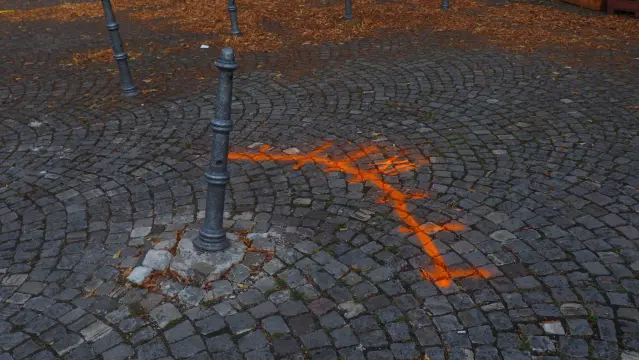 Schief stehender Poller im Kopfsteinpflaster. Mit orangener Farbe ist ein Bogen und die römische Zahl 12  auf das Pflaster gesprüht worden.