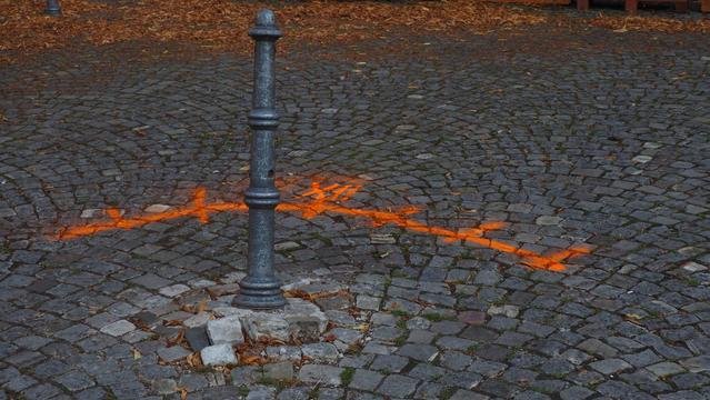 Schief stehender Poller im Kopfsteinpflaster. Mit orangener Farbe ist ein Bogen und die römische Zahl 12  auf das Pflaster gesprüht worden.
