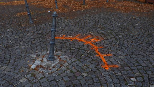 Schief stehender Poller im Kopfsteinpflaster. Mit orangener Farbe ist ein Bogen und die römische Zahl 12  auf das Pflaster gesprüht worden.