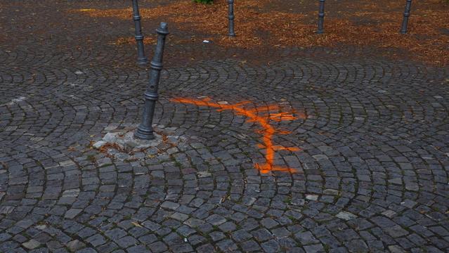 Schief stehender Poller im Kopfsteinpflaster. Mit orangener Farbe ist ein Bogen und die römische Zahl 12  auf das Pflaster gesprüht worden.