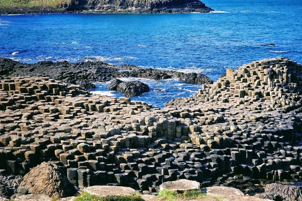 A wide shot of the Giant's Causeway in Northern Ireland, showcasing the distinctive hexagonal basalt columns in various sizes along the coastline. The deep blue sea stretches out behind the columns, with rocky cliffs visible in the distance under a bright sky.