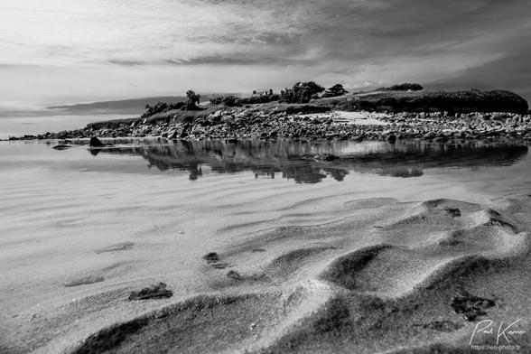 Photographie monochrome en noir et blanc, présentant un paysage capté au ras du sable d'une plage, juste devant une flaque d'eau où se reflètent  les rochers du rivage et la végétation d'une île dominée par la toiture d'une maison entre des arbres. Le bas de la photo est constitué des ondulations du sable à marée basse puis, au dessus, la flaque d'eau qui semble immense du fait de sa proximité avec l'objectif alors qu'un ciel partiellement nuageux constitue le haut de la photo, l'île et son reflet dans la flaque étant entre ciel et sable.