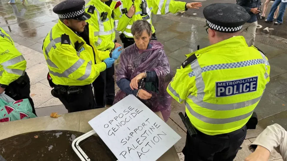 Photograph from yesterday's protest in support of Palestine Action in London.  A late middle-aged lady with short dark grey hair, wearing a plum-coloured rain poncho, is sitting on the ground, handcuffed, surrounded by police officers.  Beside her in the foreground is a placard reading I OPPOSE GENOCIDE I SUPPORT PALESTINE ACTION