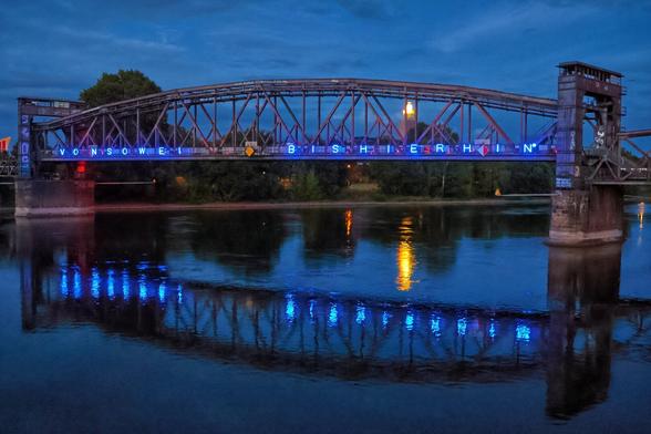 A captivating long exposure photograph at dusk showcases a large, dark metal railway bridge, Hubbrücke in Magdeburg, spanning the wide river Elbe. Blue illuminated text on the bridge,  "VON SO WEIT BIS HIER HIN", is reflected vibrantly in the still, dark blue water below. A warm, golden light emanates from a structure on the far bank, also reflecting in the water. The sky is a deep twilight blue with visible clouds. The overall scene is serene and dramatic, highlighting the bridge's architecture and the reflections on the water.