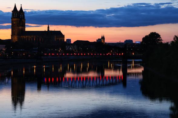 A breathtaking twilight scene captures the city of Magdeburg across a river, with the sky transitioning from a fiery orange-red near the horizon to deep blue above. The prominent silhouette of Magdeburg Cathedral, with its twin spires, stands tall on the left against the vibrant sky. The same bridge as in the previous image, Hubbrücke, but viewed from the other side, spans the river, adorned with red illuminated text that is clearly reflected in the calm water. The text reads "VON HIER AUS NOCH VIEL WEITER". The city lights twinkle along the riverbank, adding to the picturesque ambiance.
