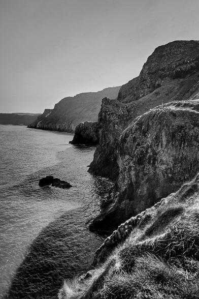 A dramatic black and white photograph captures a rugged coastline with towering cliffs descending into the calm, dark waters of the sea. The sun casts a strong light from the right, creating deep shadows and highlighting the textured rock faces and patches of sparse grass on the cliff edges. The line of cliffs recedes into the distance, with the horizon line slightly visible under a clear sky. A few small rocks are visible in the water near the base of the closest cliff.
