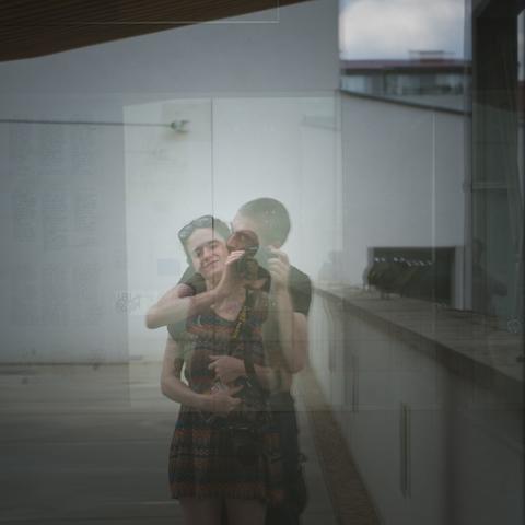 A couple make a selfie. The man hold the women around her shoulders, holding the camera in front of them. The portrait is visible in the reflection of a glass window