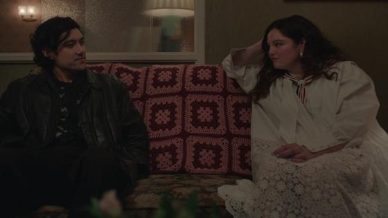 Una escena de dos personajes sentados en un sofá, uno de ellos viste de negro y el otro lleva una blusa blanca, en un ambiente acogedor con luz suave.